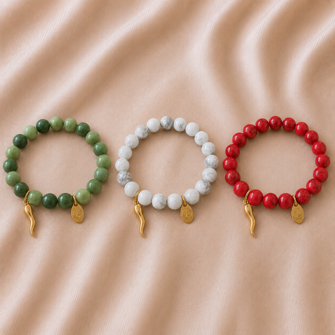 Three beaded bracelets arranged in a row on a soft beige fabric background. The left bracelet is green, the center bracelet is white and gray marbled, and the right bracelet is red, each with a small gold charm at the bottom.