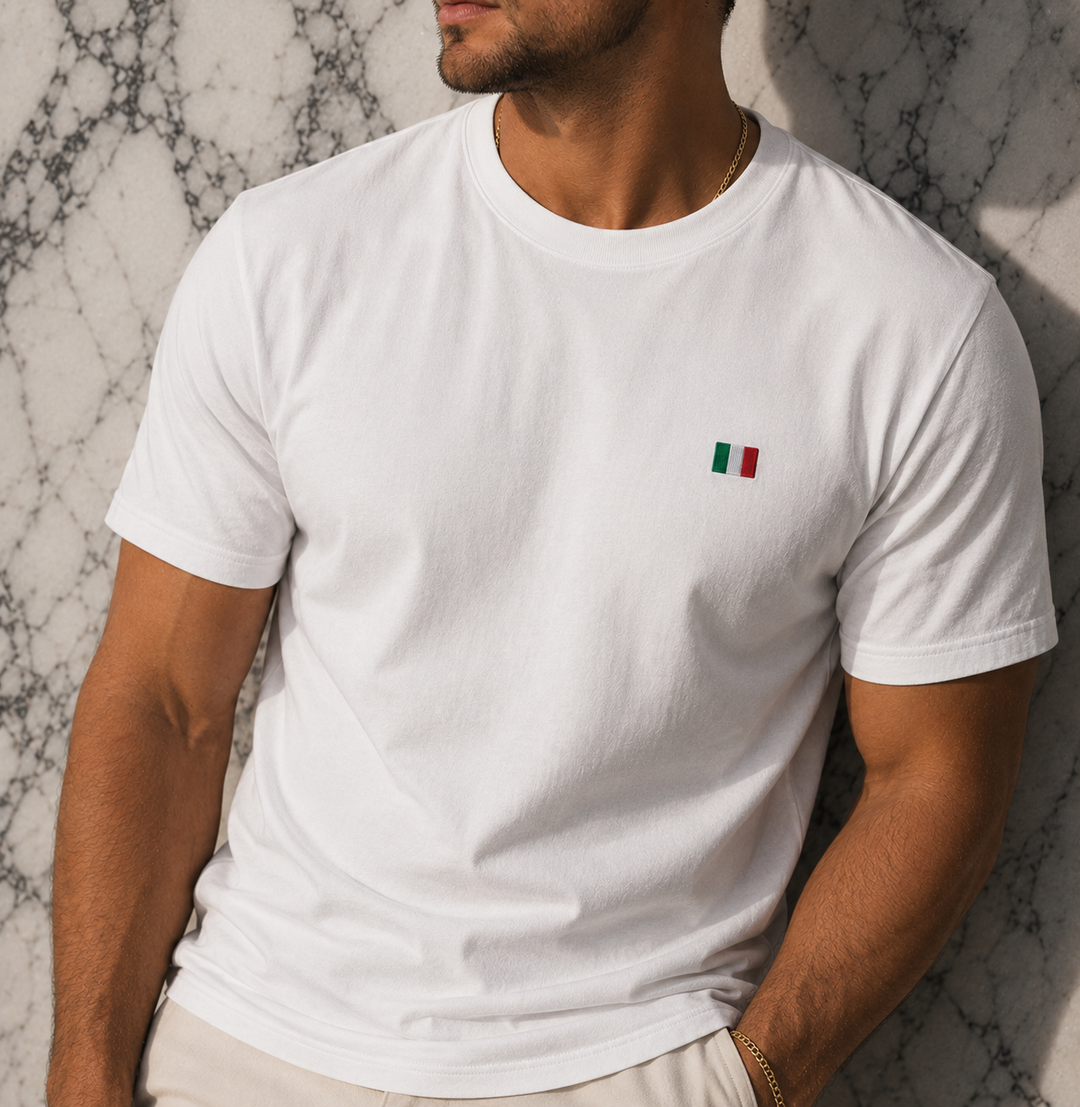 Person wearing a white t-shirt with a small flag emblem against a marble wall.